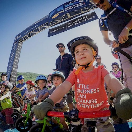 Children with helmets on their bikes at a starting line for a race, one in a red shirt that reads 