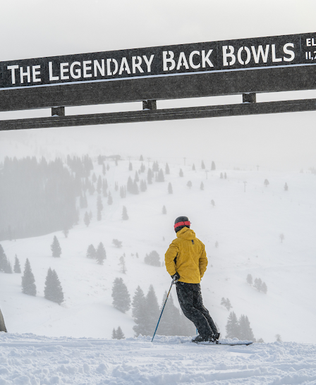 A skier in a yellow jacket stands under a sign reading "The Legendary Back Bowls Elev. 11,250" with a snowy mountain landscape and trees in the background.