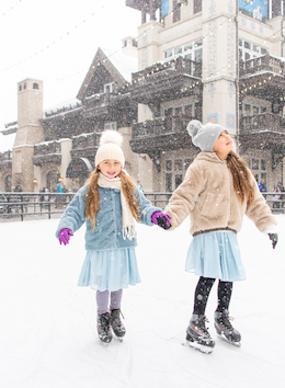 Two children are ice skating outdoors, holding hands, and smiling. They are dressed warmly in winter outfits, and it's snowing.