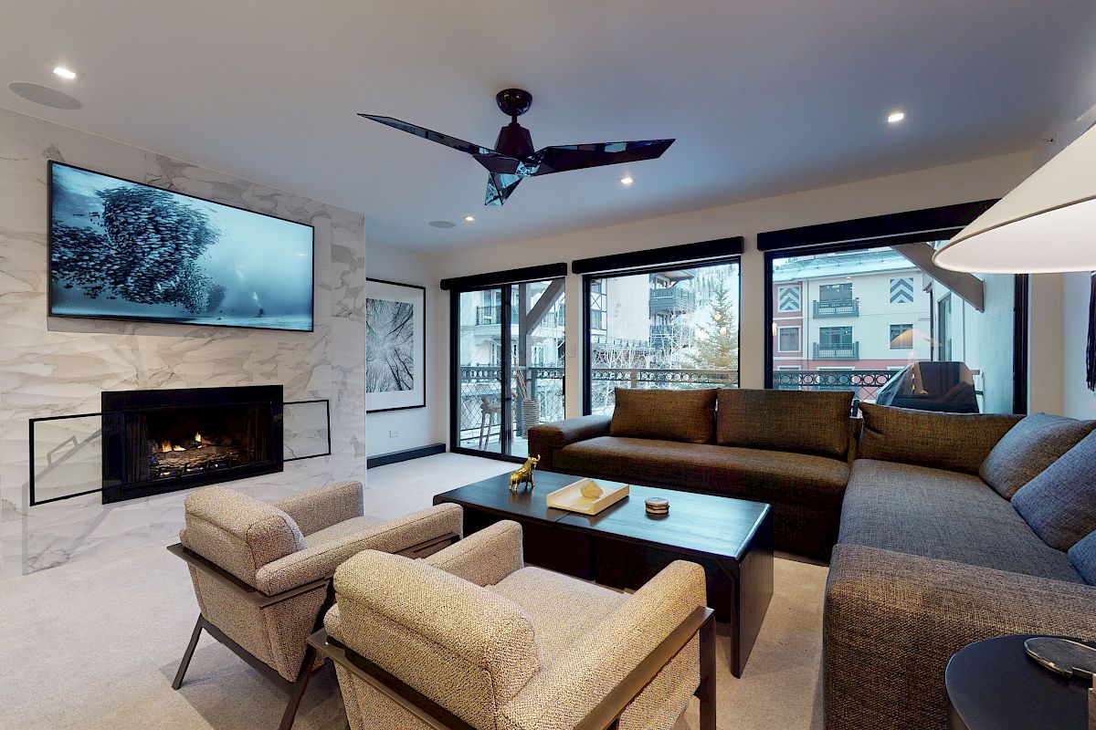 A modern living room with a large sectional sofa, two armchairs, a coffee table, a wall-mounted TV, and a fireplace, with large windows.