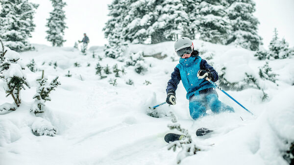 A person is skiing down a snow-covered slope, surrounded by trees and wearing a helmet and ski gear, with another person in the background.