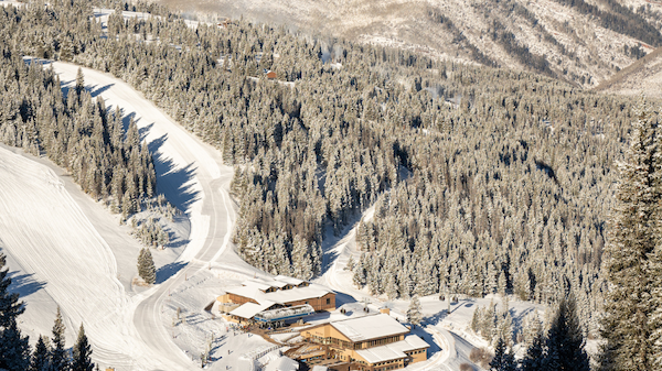 A snowy mountain resort nestled among pine trees with ski slopes and several buildings at the base amidst a vast forest landscape.