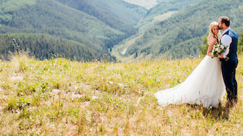 A couple in wedding attire kisses on a grassy hilltop with mountains and valleys in the background, under a clear sky.