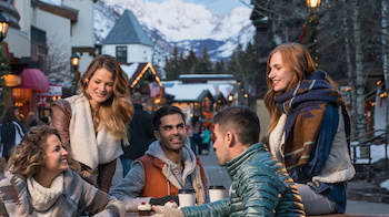 A group of people dressed in winter clothing are sitting at an outdoor table in a picturesque snowy village with mountains in the background.