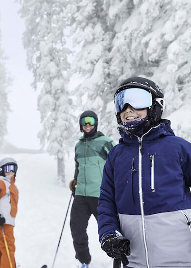 A group of three people, dressed in winter gear and ski helmets, stand on a snowy slope surrounded by snow-covered trees, presumably enjoying skiing.