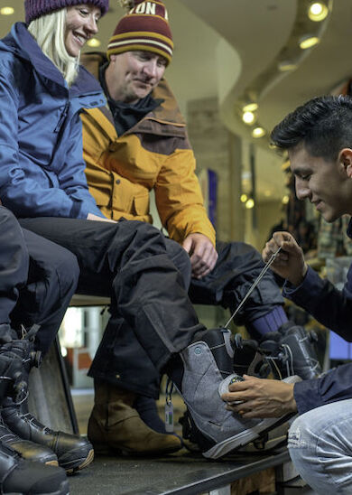 A person is assisting another with fitting snowboard boots while two others watch in a store setting, all dressed in winter sports gear.