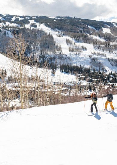 Three people are skiing on a snowy slope with a scenic view of mountains and a town in the background, under a partly cloudy sky.