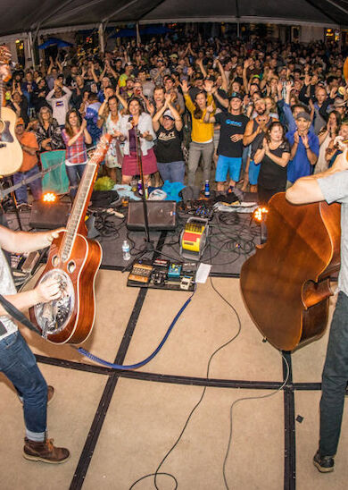 A band performs on stage, viewed from behind, with a lively audience cheering and clapping under a tented venue.