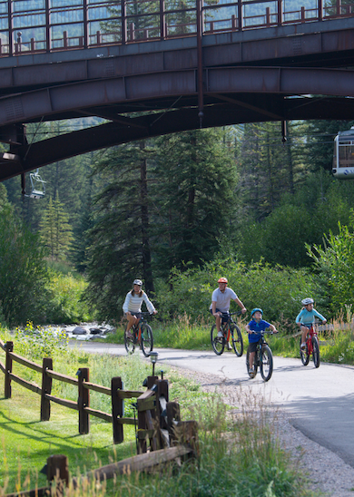 A family rides bicycles on a paved path under a bridge, surrounded by green trees and a scenic landscape, with a cable car visible in the background.