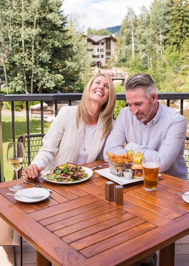 A couple is sitting at an outdoor table enjoying a meal with drinks, smiling and surrounded by a scenic view with trees and buildings in the background.