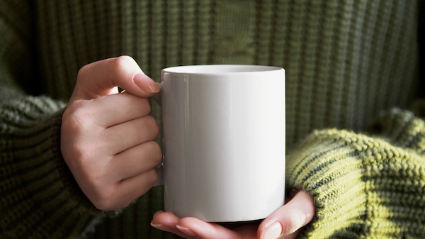 A person in a green sweater is holding a plain white mug with both hands.