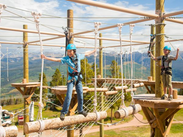 Two children wearing helmets and harnesses navigate a ropes course outdoors, with a scenic mountain backdrop. They appear to be enjoying the activity.