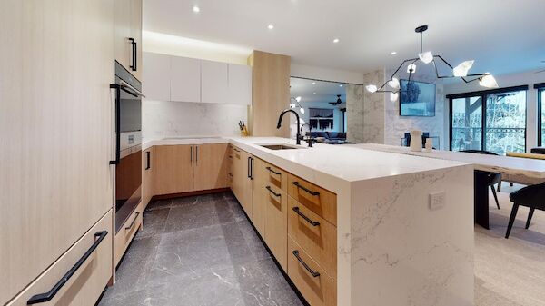 A modern kitchen featuring light wood cabinets, a large island with a sink, marble countertops, dark hardware, and a contemporary chandelier.