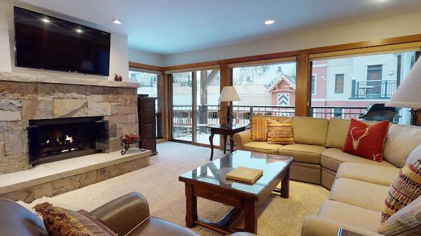 A cozy living room with a stone fireplace, large sectional sofa, wooden coffee table, and big windows overlooking a balcony and building.