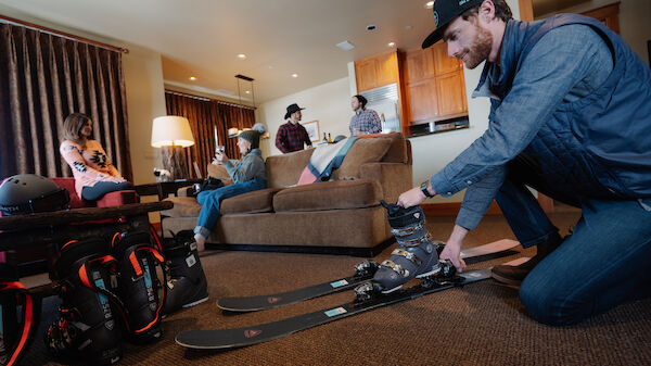 A group of people is in a cozy living room preparing skiing gear, with one person adjusting ski bindings and others seated on the couch.