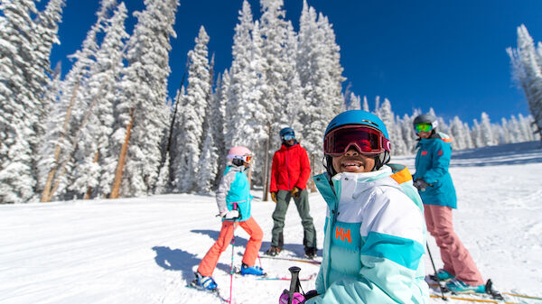 A group of people in colorful ski gear are on a snowy slope with tall, frosty trees in the background, smiling under a clear blue sky.