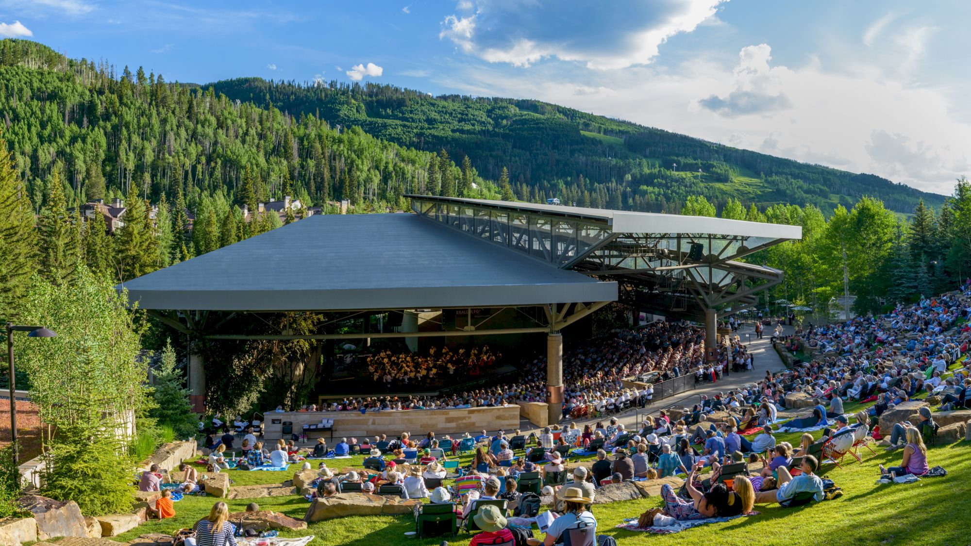 An outdoor concert with a large audience on a grassy slope, surrounded by trees and scenic mountains. The sky is partly cloudy.