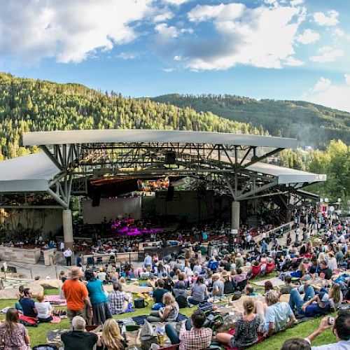 An outdoor concert venue surrounded by mountains and trees, with a large crowd seated on grass and under a roof, enjoying a live performance.