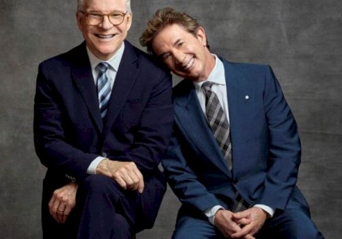 Two men in suits are posing for a photo; one is sitting with his hands clasped, and the other is leaning his head on the man's shoulder, both smiling.