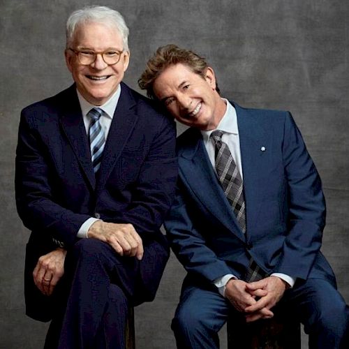 Two men in suits are posing for a photo; one is sitting with his hands clasped, and the other is leaning his head on the man's shoulder, both smiling.