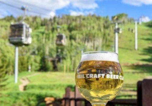 A glass of craft beer sits on a ledge with a view of a green hillside and ski lifts in the background under a partly cloudy sky, ending the sentence.