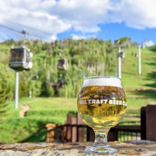 A glass of craft beer sits on a ledge with a view of a green hillside and ski lifts in the background under a partly cloudy sky, ending the sentence.