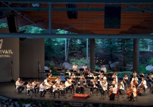 An orchestra is performing on stage at the Bravo! Vail Music Festival, with musicians playing various instruments. The backdrop features a natural forest scene.