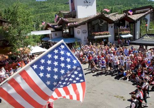 A festive outdoor gathering with a large American flag in the foreground and a crowd of people near a building with alpine architecture.