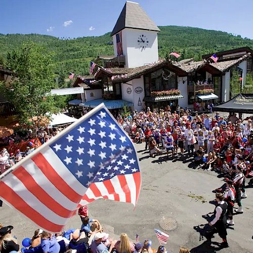 A festive outdoor gathering with a large American flag in the foreground and a crowd of people near a building with alpine architecture.