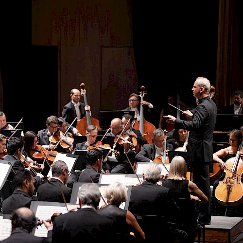 A conductor leads an orchestra during a performance; musicians play various instruments, with 