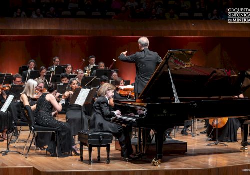 A symphony orchestra performs on stage with a conductor leading and a pianist playing a grand piano, in front of the Orchestra Sinfonica Mineria.