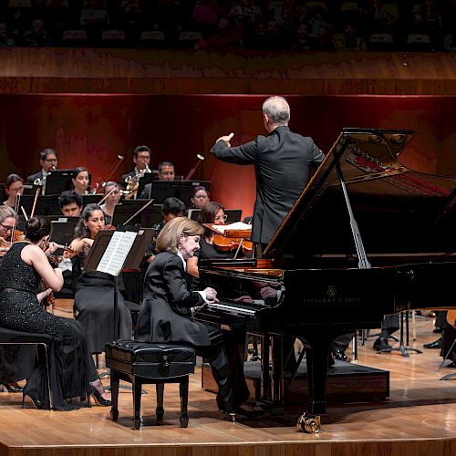 A symphony orchestra performs on stage with a conductor leading and a pianist playing a grand piano, in front of the Orchestra Sinfonica Mineria.