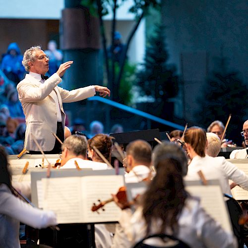 An orchestra conductor leads a group of musicians during a performance, with an audience in the background.