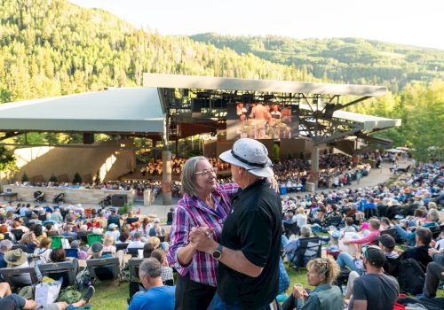 A couple dances in the foreground while a large crowd sits on the grass watching a performance at a scenic outdoor amphitheater in the mountains.