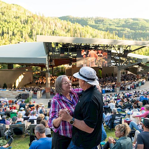 A couple dances in the foreground while a large crowd sits on the grass watching a performance at a scenic outdoor amphitheater in the mountains.