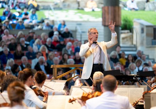 An orchestra performs with a conductor leading, surrounded by a seated audience in an outdoor venue.