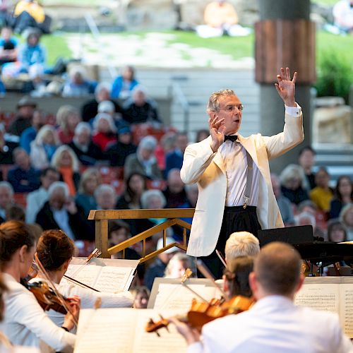 An orchestra performs with a conductor leading, surrounded by a seated audience in an outdoor venue.