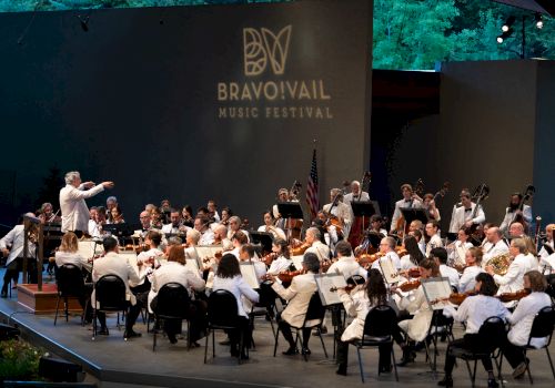 An orchestra performs at the Bravo! Vail Music Festival with a conductor leading the musicians, all dressed in white, under a night sky.