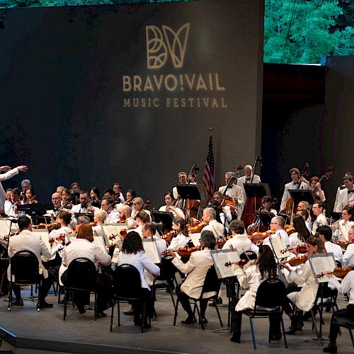 An orchestra performs at the Bravo! Vail Music Festival with a conductor leading the musicians, all dressed in white, under a night sky.