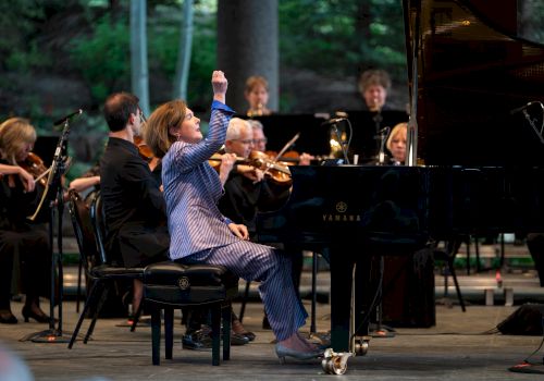 A pianist performs passionately on stage, surrounded by an orchestra, raising one arm while playing the piano, with musicians focused on their instruments.