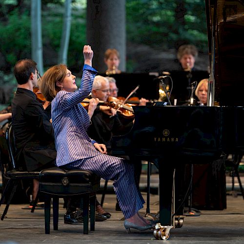 A pianist performs passionately on stage, surrounded by an orchestra, raising one arm while playing the piano, with musicians focused on their instruments.