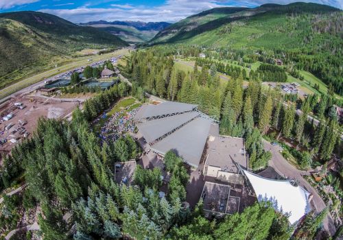 An aerial view of a large building complex surrounded by dense forested mountains, with a valley and road visible in the distance.