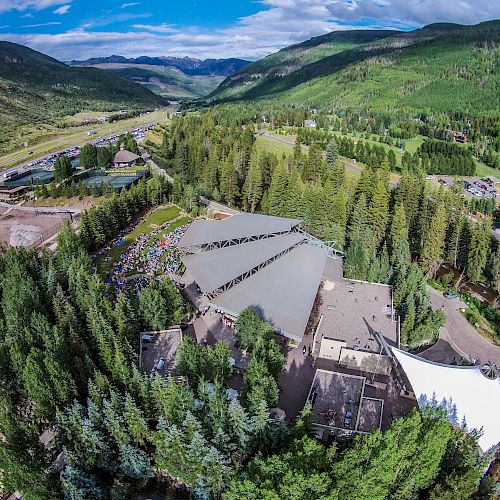 An aerial view of a large building complex surrounded by dense forested mountains, with a valley and road visible in the distance.