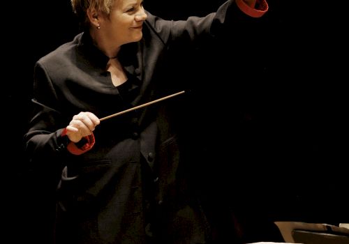 The image features a person in a dark suit conducting an orchestra with a baton, standing behind a music stand with sheet music on it.