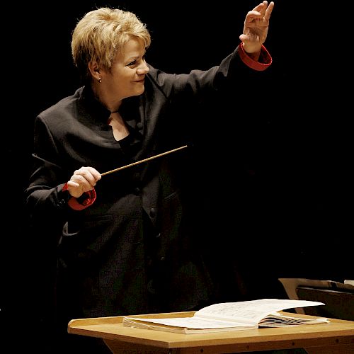 The image features a person in a dark suit conducting an orchestra with a baton, standing behind a music stand with sheet music on it.