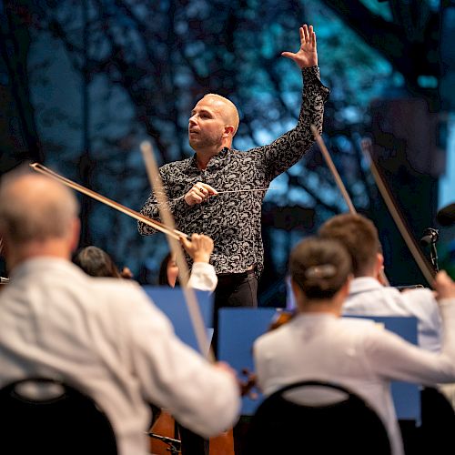 A conductor leading an orchestra performance, with musicians playing string instruments, and the background showcasing a serene, tree-filled scene.