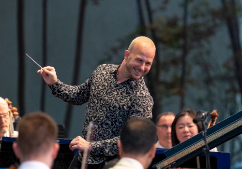 A conductor leads an orchestra during a performance, with musicians playing various instruments and reading sheet music. Trees are visible in the background.