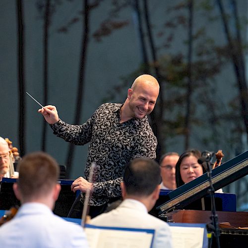 A conductor leads an orchestra during a performance, with musicians playing various instruments and reading sheet music. Trees are visible in the background.