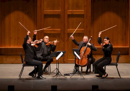 A string quartet dressed in black performs on stage with wooden paneling, raising their bows in unison after playing a piece.