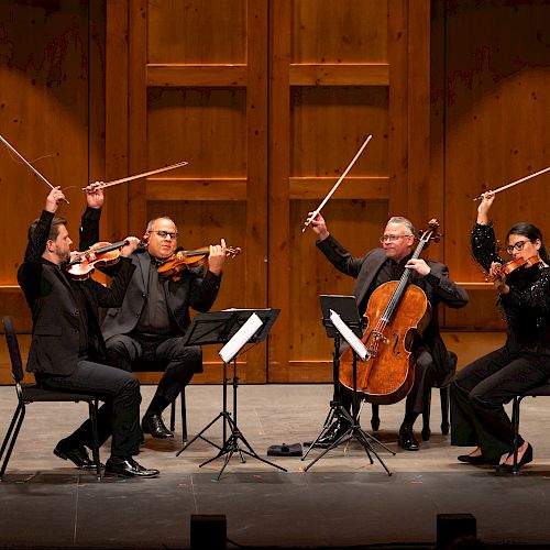 A string quartet dressed in black performs on stage with wooden paneling, raising their bows in unison after playing a piece.